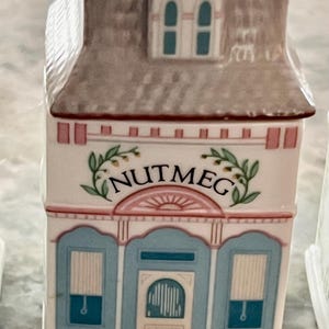 May include: A white ceramic spice jar shaped like a house with a pink roof and blue trim. The jar has a pink and white sign that says "NUTMEG" with green leaves on either side.