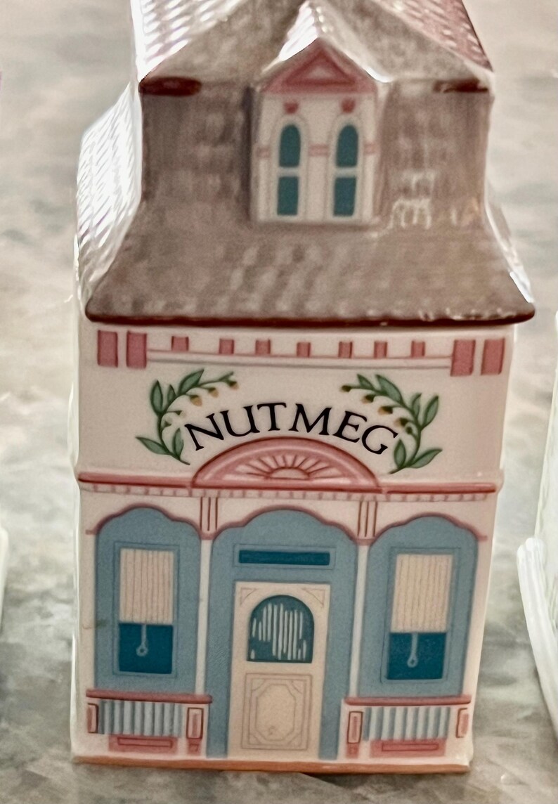 May include: A white ceramic spice jar shaped like a house with a pink roof and blue trim. The jar has a pink and white sign that says "NUTMEG" with green leaves on either side.