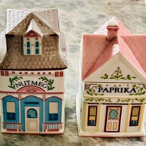 May include: Two ceramic spice jars shaped like houses. The jar on the left is brown with blue trim and says "Nutmeg". The jar on the right is pink with yellow trim and says "Paprika".
