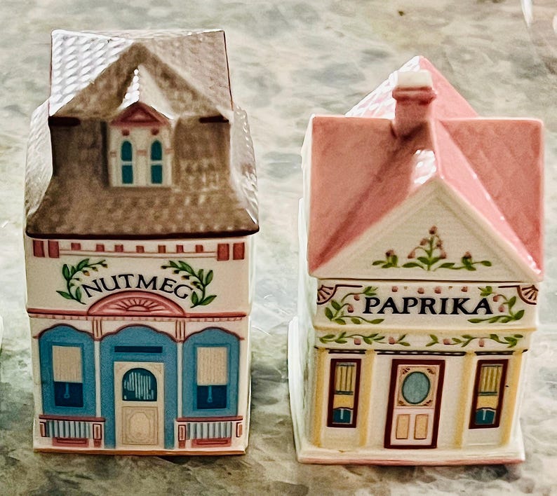 May include: Two ceramic spice jars shaped like houses. The jar on the left is brown with blue trim and says "Nutmeg". The jar on the right is pink with yellow trim and says "Paprika".