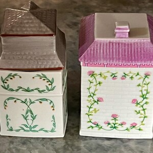May include: Set of three ceramic canisters with floral designs. The canisters are shaped like houses with pink, white and grey roofs. The canisters have a white background with green vines and pink flowers.