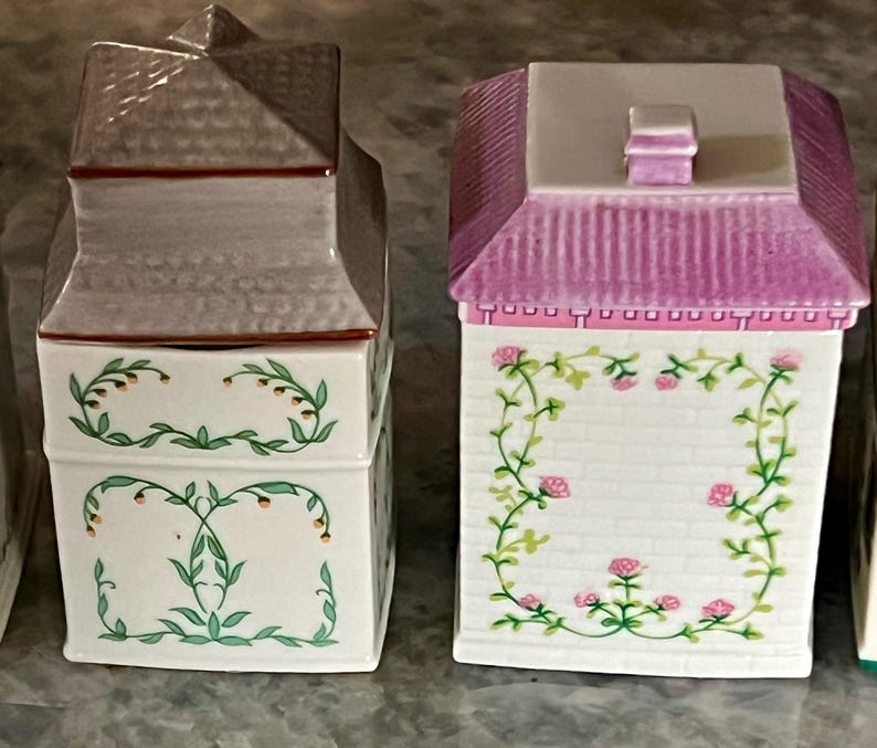 May include: Set of three ceramic canisters with floral designs. The canisters are shaped like houses with pink, white and grey roofs. The canisters have a white background with green vines and pink flowers.
