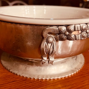 May include: A silver bowl with a white porcelain interior and decorative floral accents. The bowl has a scalloped edge and sits on a pedestal base.
