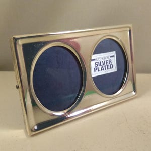 May include: Double photo frame with two round openings for pictures, silver-plated. The frame has a label that reads "Genuine Silver Plated".