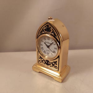 Può includere: Un orologio da tavolo in metallo dorato con parte superiore ad arco e quadrante circolare bianco. L'orologio presenta numeri romani neri, lancette nere e la scritta Le Temps Quartz. Sono visibili intagli decorativi in filigrana nera.