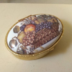 Op de afbeelding: Een klein ovaal sieradendoosje met een gouden rand en een wit porseleinen deksel. Het deksel heeft een kleurrijke illustratie van een mand vol fruit, waaronder druiven, appels, peren en pruimen.