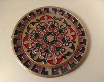 May include: A round, brass plate with a colourful, geometric design. The design features a central flower with red, green, and blue accents. The plate is surrounded by a black and brown border.