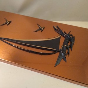 Op de afbeelding: Een koperkleurig rechthoekig wandkunstwerk met een zeilboot- en vogelontwerp. De zeilboot is zwart en goud, en de vogels zijn donker. Het kunstwerk heeft een metallic afwerking en is gemonteerd op een wit oppervlak.