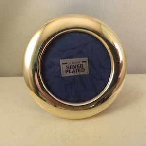 May include: A gold-toned, round picture frame with a blue velvet backing. The frame has a raised edge and says "GENUINE SILVER PLATED" on the front.
