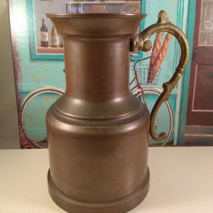 Può includere: Un'anfora di rame con un manico decorativo. L'anfora ha un corpo arrotondato e una bocca larga.