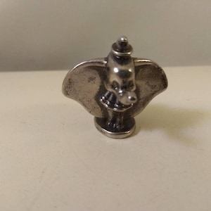 Pode incluir: Pequena figura prateada do elefante Dumbo, com orelhas grandes e um design detalhado. A figura tem uma pequena base e é provavelmente um item de colecionador ou decorativo.