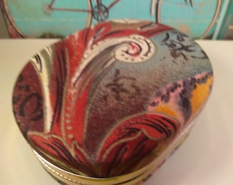 May include: A round, vintage trinket box with a gold metal rim. The box is covered in a colorful fabric with a swirling, abstract pattern in shades of red, brown, blue, and yellow.
