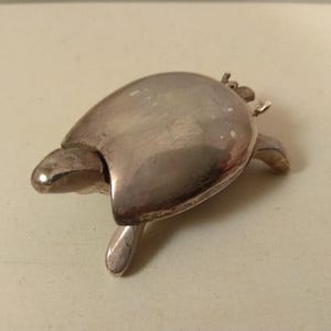 以下が含まれることがあります： 銀色のカメの形をした小物入れ。箱は丸みを帯びた甲羅と4本の小さな脚があります。カメの頭は伸びており、箱は前面で開くようです。カメは光沢のある反射面を持っています。