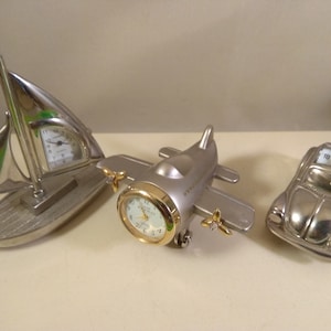 Może przedstawiać: Trzy srebrne miniaturowe zegary w kształcie żaglówki, samolotu i samochodu. Każdy zegar ma białą tarczę z czarnymi cyframi i wskazówkami.