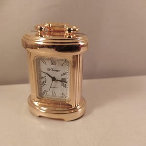 Può includere: Un piccolo orologio da carrozza color oro con finitura metallica lucida. Il quadrante bianco rettangolare presenta numeri romani neri, lancette nere e la scritta Le Temps e Quartz stampata in nero. Dotato di una maniglia decorativa superiore.