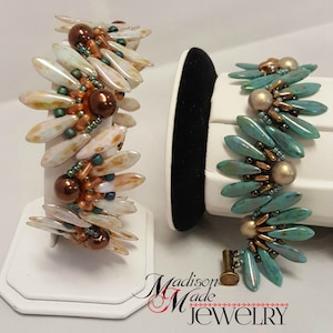 Könnte beinhalten: Zwei Perlenarmbänder mit einem einzigartigen Design. Ein Armband ist aus weißen und braunen Perlen gefertigt, das andere aus türkisfarbenen und goldenen Perlen. Beide Armbänder werden auf weißen Ständern präsentiert.