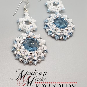 May include: A pair of silver earrings with blue and white crystal beads. The earrings feature a larger round bead with a blue crystal in the center, surrounded by smaller white and silver beads. The earrings are attached to silver hooks.