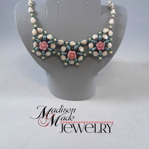 May include: A white and green beaded necklace with three pink flower accents. The necklace is displayed on a gray mannequin bust. The text "Madison Made Jewelry" is visible below the bust.