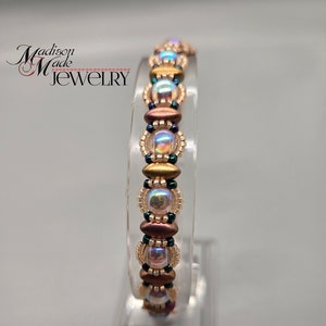 Puede incluir: Un brazalete de cuentas con cuentas iridiscentes, doradas y marrones. El brazalete se muestra en un soporte transparente.