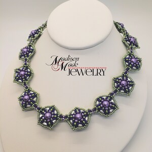 May include: A beaded necklace with a geometric design featuring purple and green beads. The necklace is on a white display stand. The text "Madison Made Jewelry" is visible in the background.