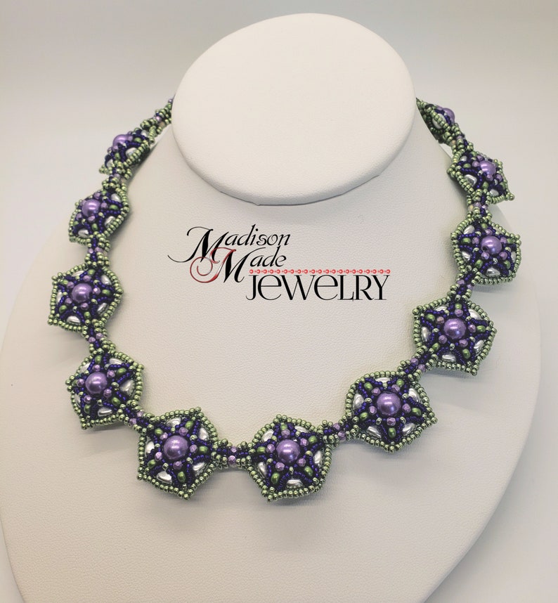 May include: A beaded necklace with a geometric design featuring purple and green beads. The necklace is on a white display stand. The text "Madison Made Jewelry" is visible in the background.