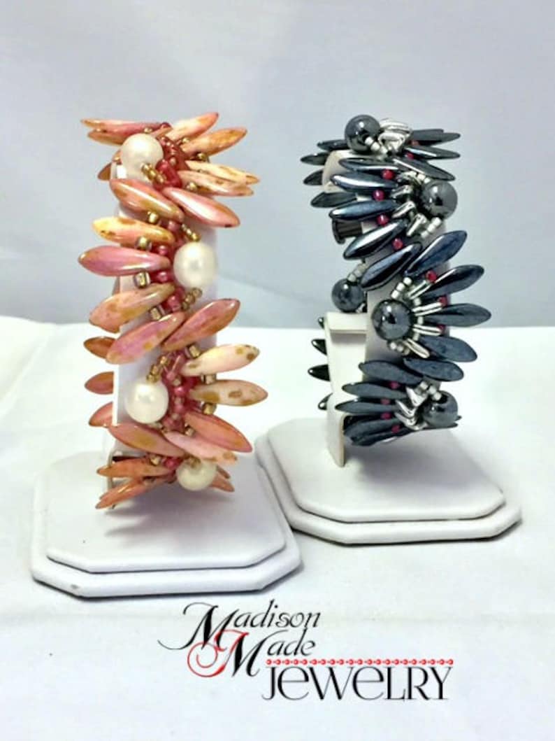 May include: Two bracelets made with beads. One bracelet is made with pink and white beads and the other is made with black and red beads. Both bracelets are on white display stands. The text "Madison Made Jewelry" is at the bottom of the image.