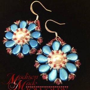 May include: A pair of blue and silver flower-shaped earrings with a white pearl in the center. The earrings are made with small beads and have a silver hook closure. The earrings are on a black background with the text "Madison Made Jewelry" in red.