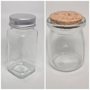 May include: Two clear glass jars. One is rectangular with a silver metal lid. The other is round with a cork stopper. Both jars are empty and suitable for storage or display of small items.