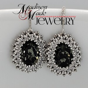 May include: A pair of silver dangle earrings with a dark gray crystal in the center of each earring. The crystal is surrounded by a silver beaded border.