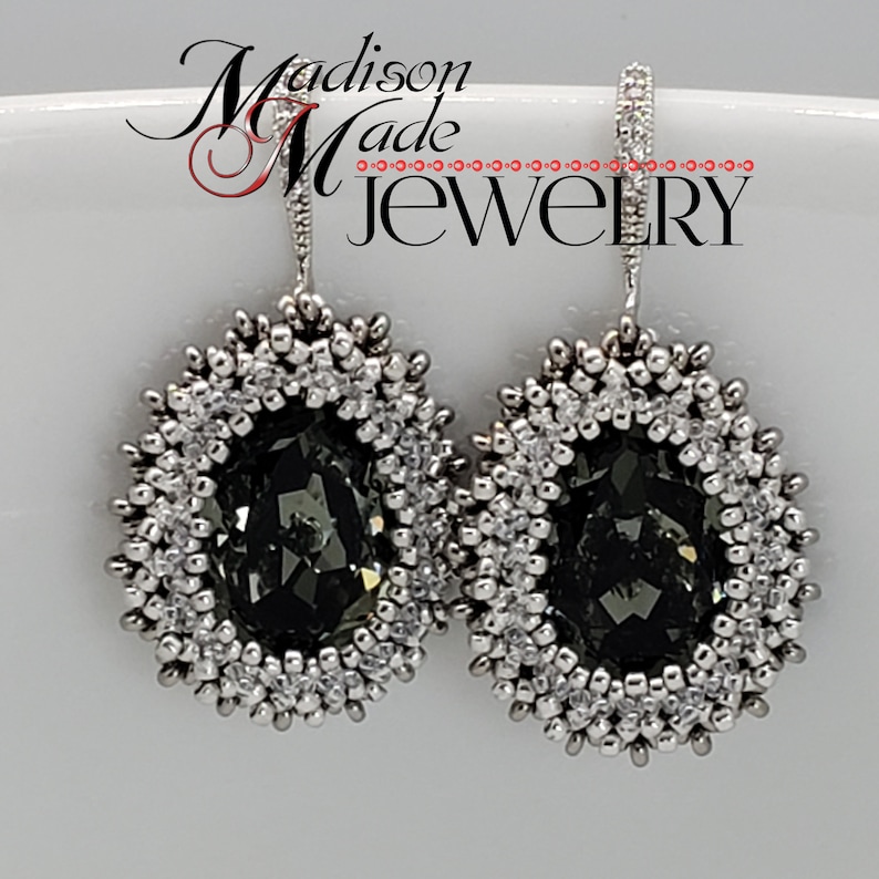 May include: A pair of silver dangle earrings with a dark gray crystal in the center of each earring. The crystal is surrounded by a silver beaded border.