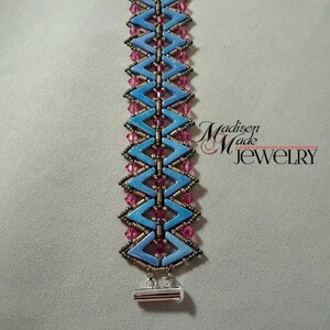 May include: A blue and pink beaded bracelet with a silver toggle clasp. The bracelet is made with small, faceted beads and has a chevron pattern.