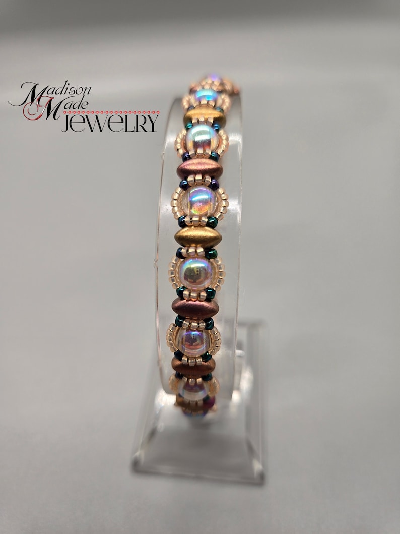 May include: A beaded bracelet with iridescent, gold, and brown beads. The bracelet is displayed on a clear stand.