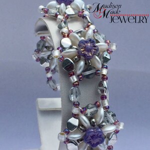 May include: A white and purple beaded bracelet with a floral design. The bracelet is made with small, round beads and features a delicate, feminine style. The bracelet is displayed on a white stand with the text "Madison Made Jewelry" in red and black.