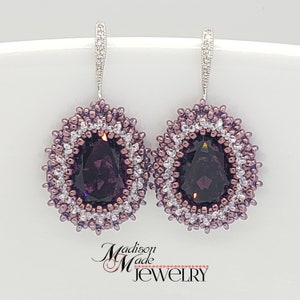 May include: A pair of dangle earrings with a large oval-shaped purple gemstone surrounded by a beaded border of silver and purple beads. The earrings have silver hooks.