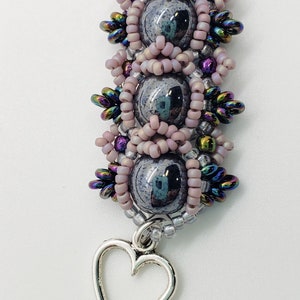 May include: A silver heart charm hangs from a beaded bracelet with pink, silver, and iridescent beads. The bracelet is made with a dark gray stone and is labeled "Hematite" and "Madison Made Jewelry".