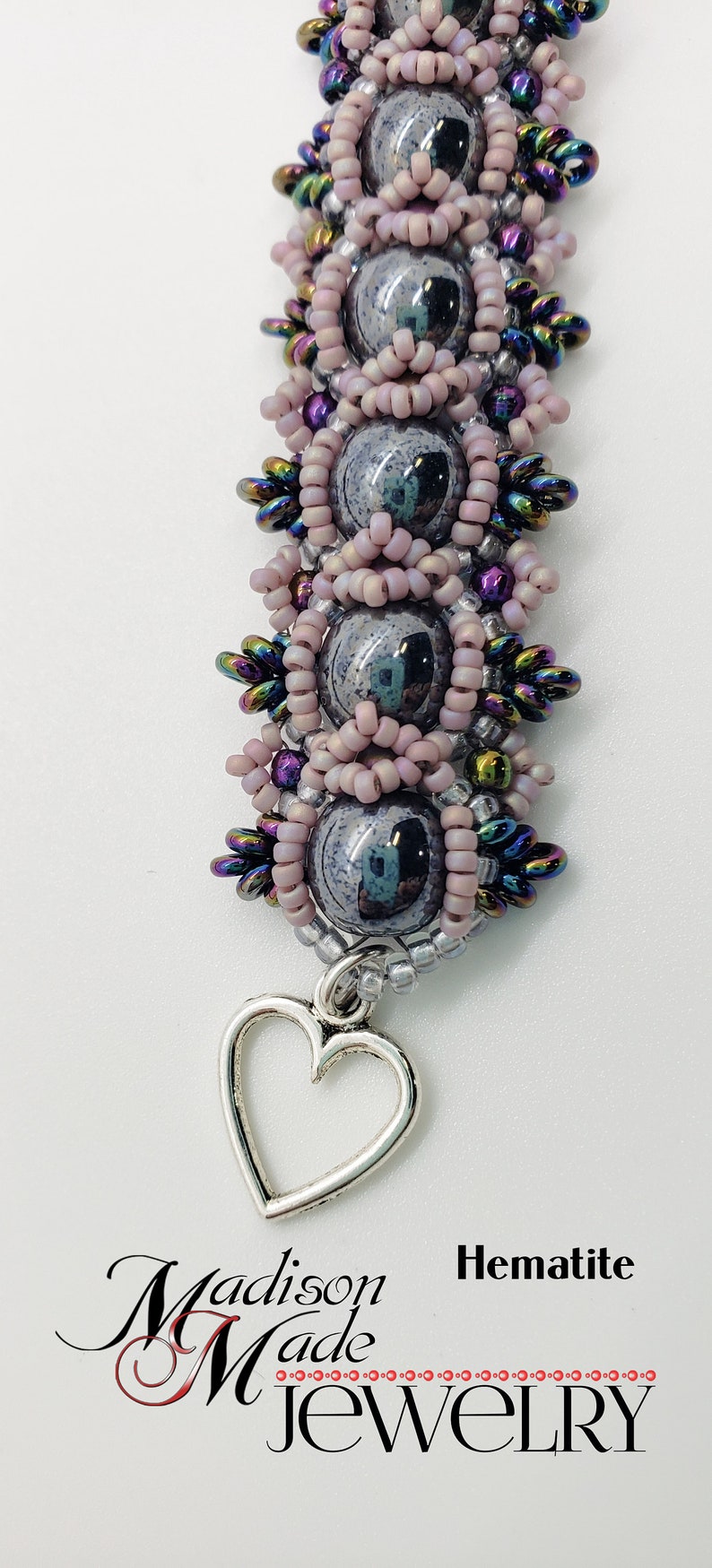 May include: A silver heart charm hangs from a beaded bracelet with pink, silver, and iridescent beads. The bracelet is made with a dark gray stone and is labeled "Hematite" and "Madison Made Jewelry".