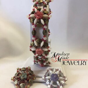 May include: A beaded bracelet with a floral design in shades of pink, white, and gold. The bracelet is displayed on a white stand with two smaller beaded flowers in different colors.