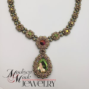 Op de afbeelding: Een groene en gouden kralenketting met een grote, gefacetteerde, druppelvormige kristallen hanger. De ketting is gemaakt met kleine, ronde kralen en heeft een herhalend patroon van kleine, ronde kristallen. De hanger is omgeven door kleinere, ronde kralen en kristallen. De ketting is gelabeld met "Madison Made Jewelry".