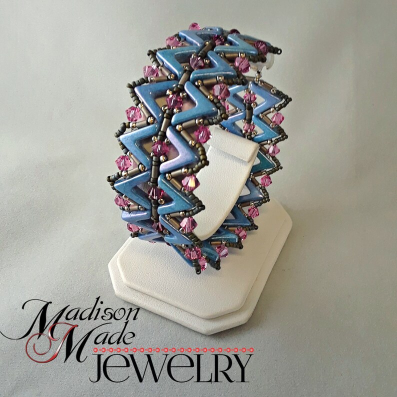 May include: A blue and pink beaded bracelet with a chevron pattern. The bracelet is on a white display stand. The text "Madison Made Jewellery" is visible in the bottom left corner of the image.