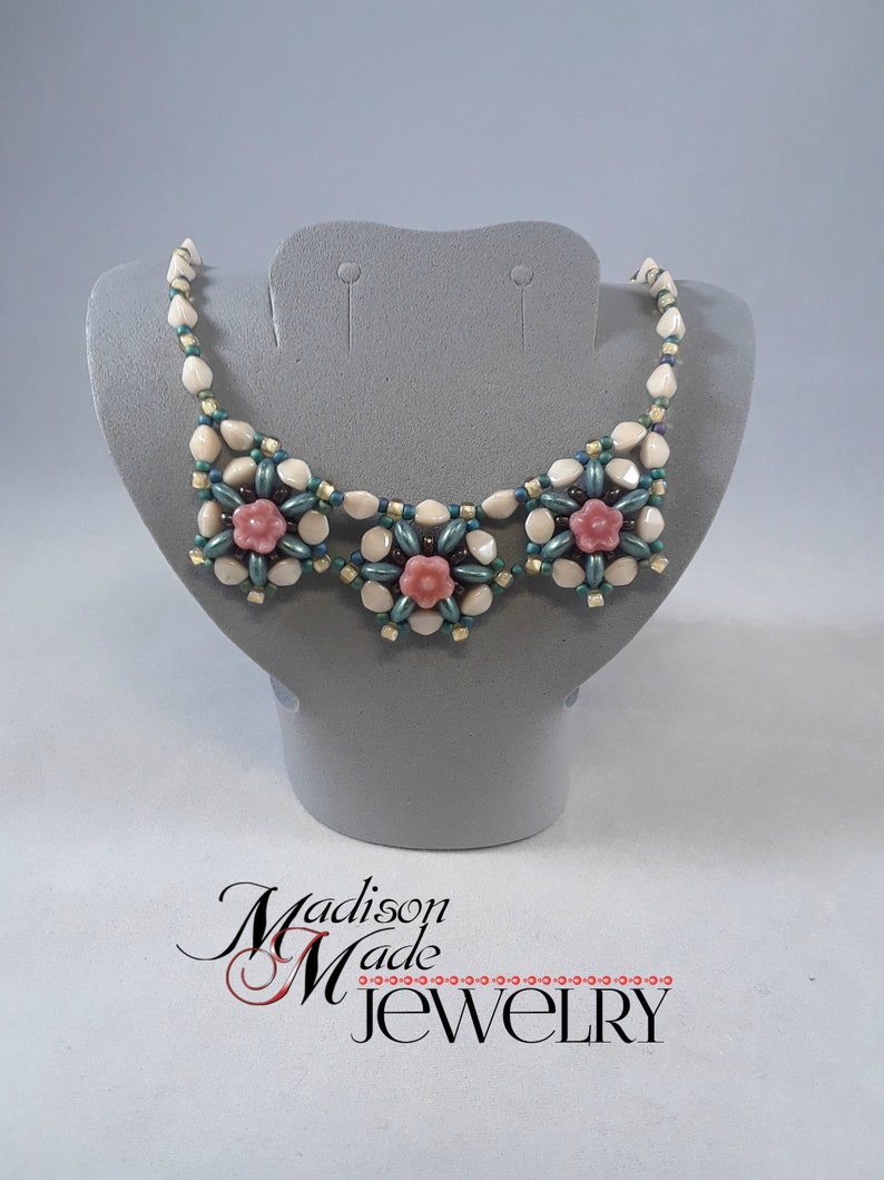 May include: A white and green beaded necklace with three pink flower accents. The necklace is displayed on a gray mannequin bust. The text "Madison Made Jewelry" is visible below the bust.