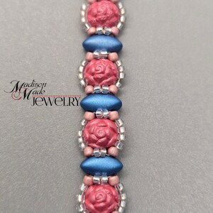 May include: A close-up of a beaded bracelet with alternating blue and pink beads. The pink beads are shaped like roses and have a textured surface. The bracelet is made with small silver beads.
