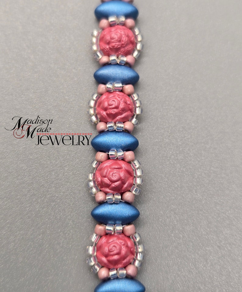 May include: A close-up of a beaded bracelet with alternating blue and pink beads. The pink beads are shaped like roses and have a textured surface. The bracelet is made with small silver beads.