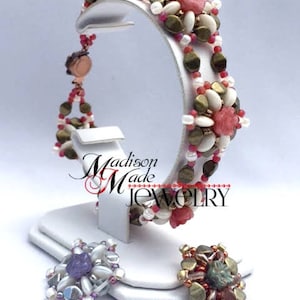 May include: A beaded bracelet with pink and white flowers, gold beads, and a brown clasp. The bracelet is on a white display stand. The text "Madison Made Jewelry" is visible on the stand.