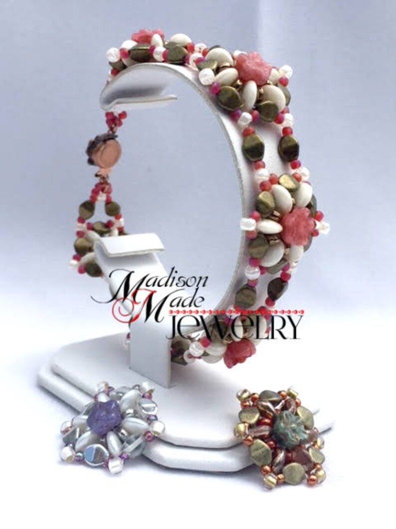 May include: A beaded bracelet with pink and white flowers, gold beads, and a brown clasp. The bracelet is on a white display stand. The text "Madison Made Jewelry" is visible on the stand.