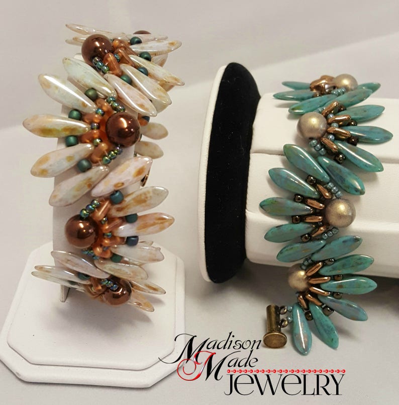 May include: Two beaded bracelets with a unique design. One bracelet is made with white and brown beads, the other is made with turquoise and gold beads. Both bracelets are displayed on white stands.