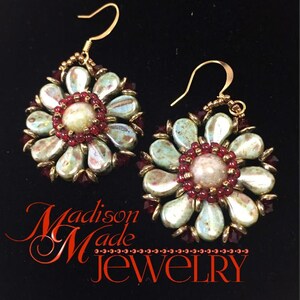 May include: A pair of gold tone earrings with a flower design. The flowers are made of iridescent green and white beads with red beads in the center. The earrings are hanging from gold hooks.