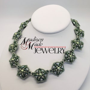 May include: A green and silver beaded necklace with a geometric design. The necklace features a repeating pattern of octagons and smaller beads. The necklace is displayed on a white mannequin.