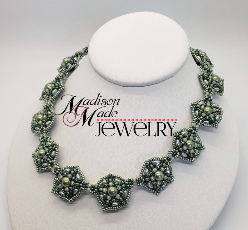 May include: A green and silver beaded necklace with a geometric design. The necklace features a repeating pattern of octagons and smaller beads. The necklace is displayed on a white mannequin.