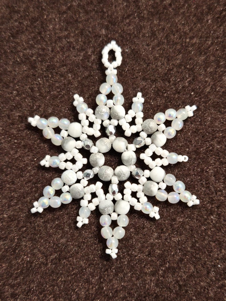 May include: A handmade snowflake ornament crafted from white and silver beads. The beaded snowflake features a starburst design with multiple points, and a loop for hanging. The ornament is displayed against a brown background.