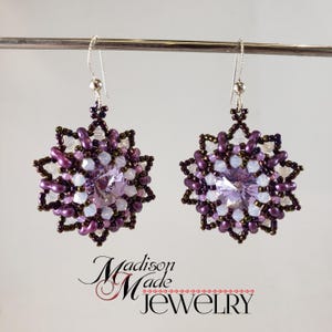 May include: A pair of silver earrings with a purple and white beaded snowflake design. The earrings feature a clear crystal in the center of each snowflake.